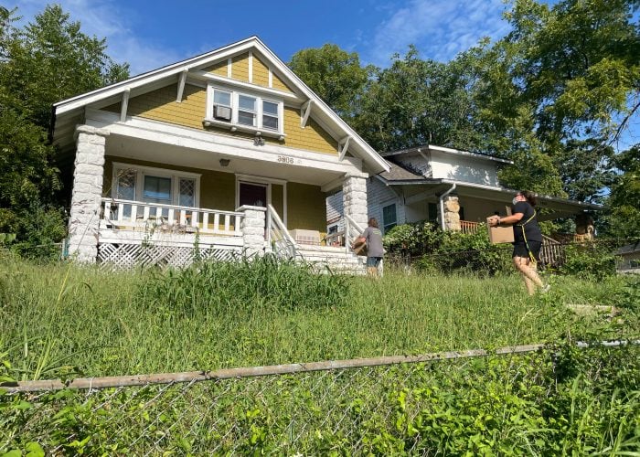 Two people walk up the sidewalk of a house with an overgrown lawn