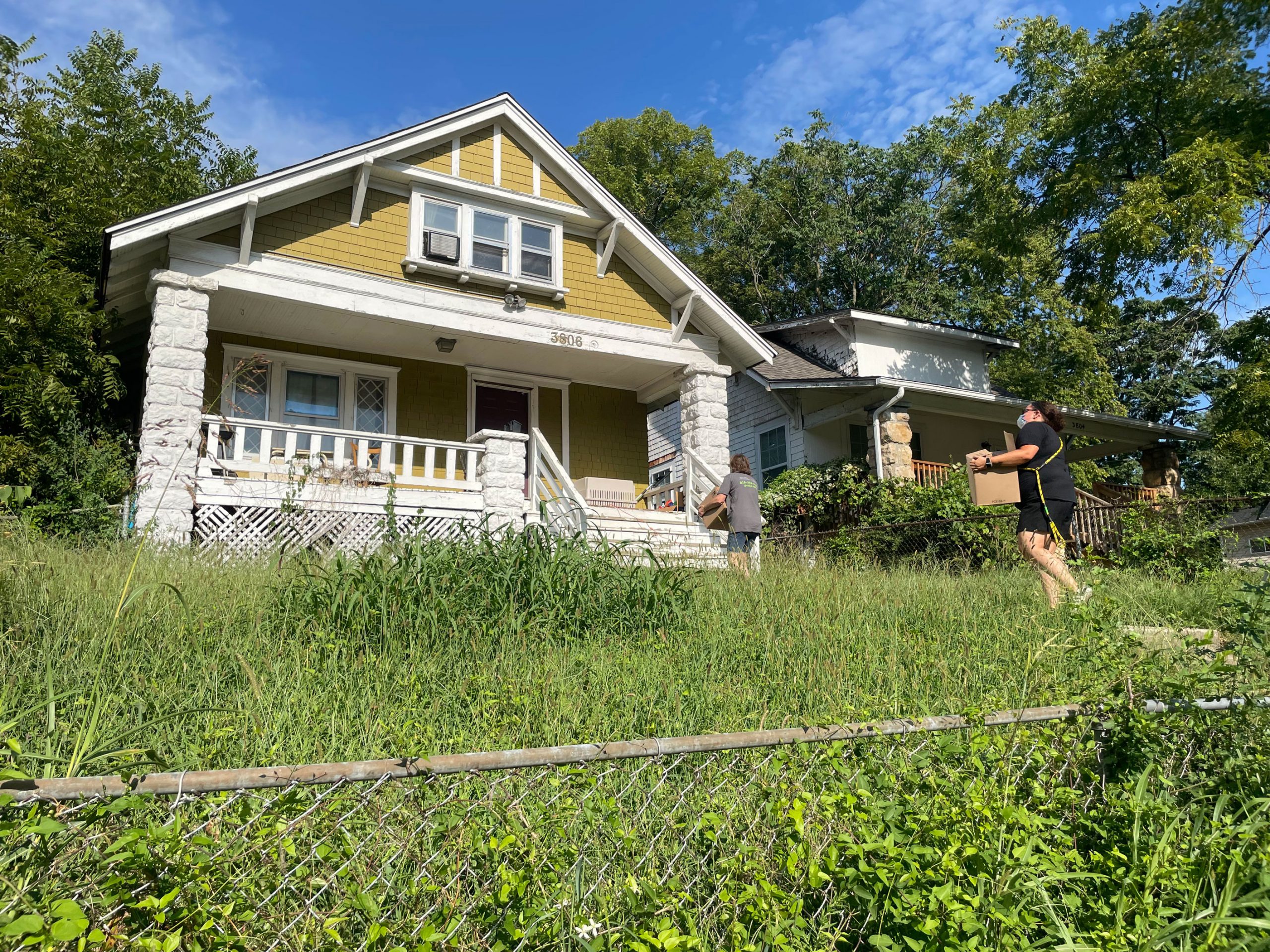 Two people walk up the sidewalk of a house with an overgrown lawn