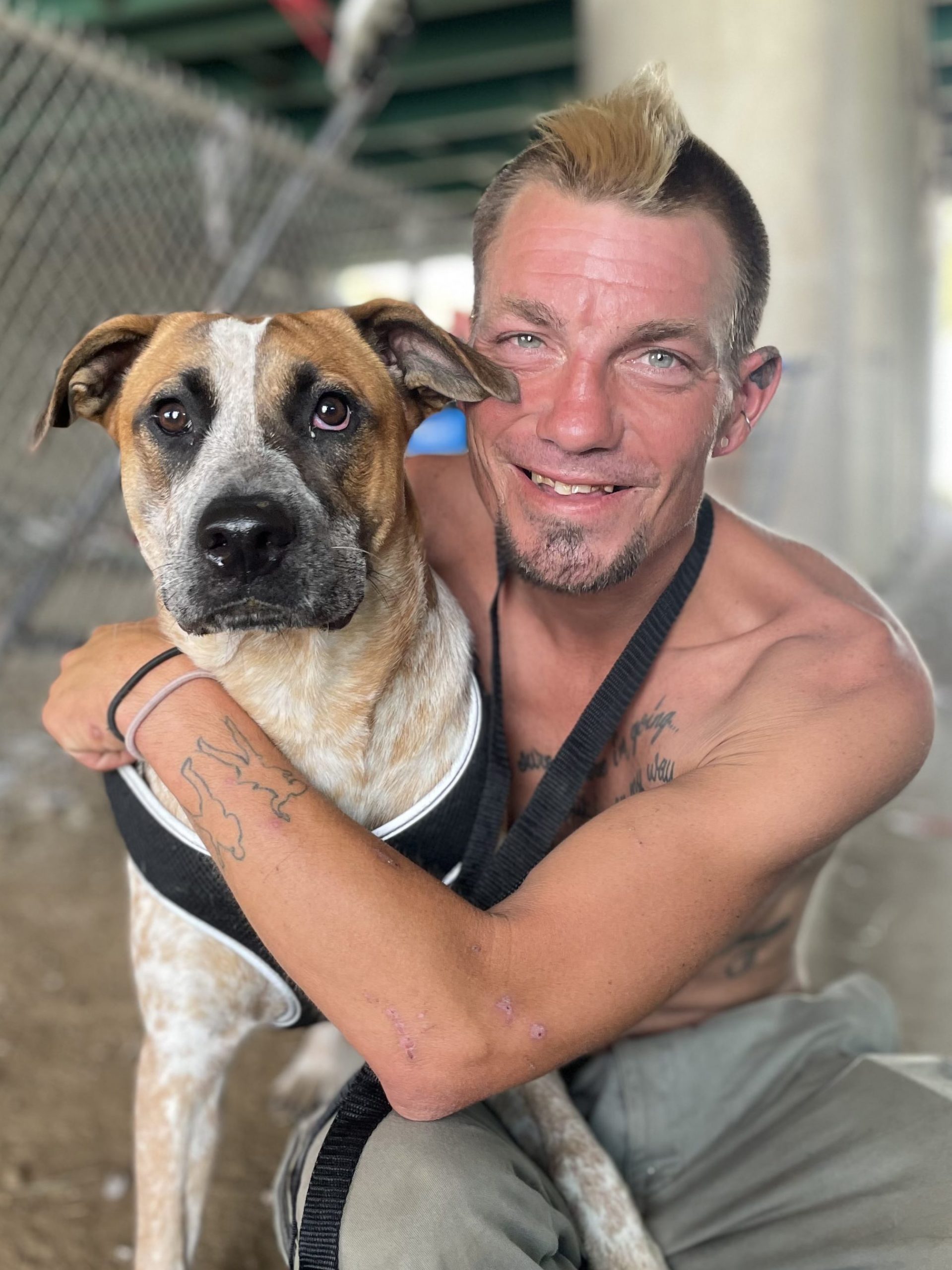 Blonde man with mohawk hugging shepherd mix underneath a bridge