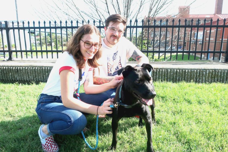 A man and woman squat and smile at the camera. Their dog looks off in a different direction.